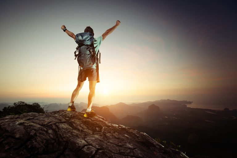 Hiker with backpack standing on top of a mountain with raised hands and enjoying sunrise